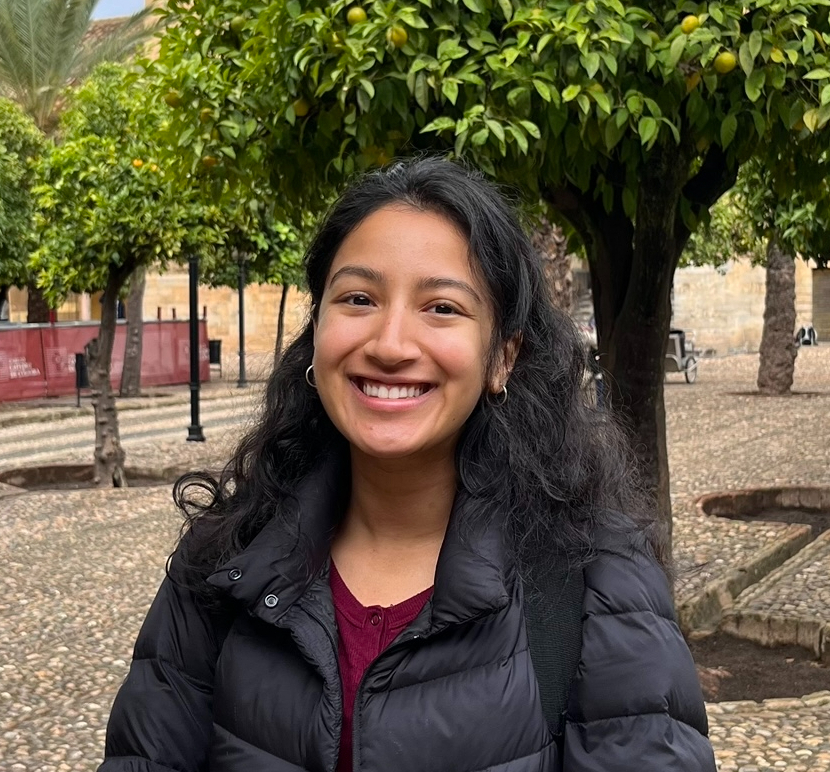 Aastha, a Nepali woman with black hair, stands in a courtyard and smiles at the camera while wearing a maroon shirt and puffy black winter jacket.